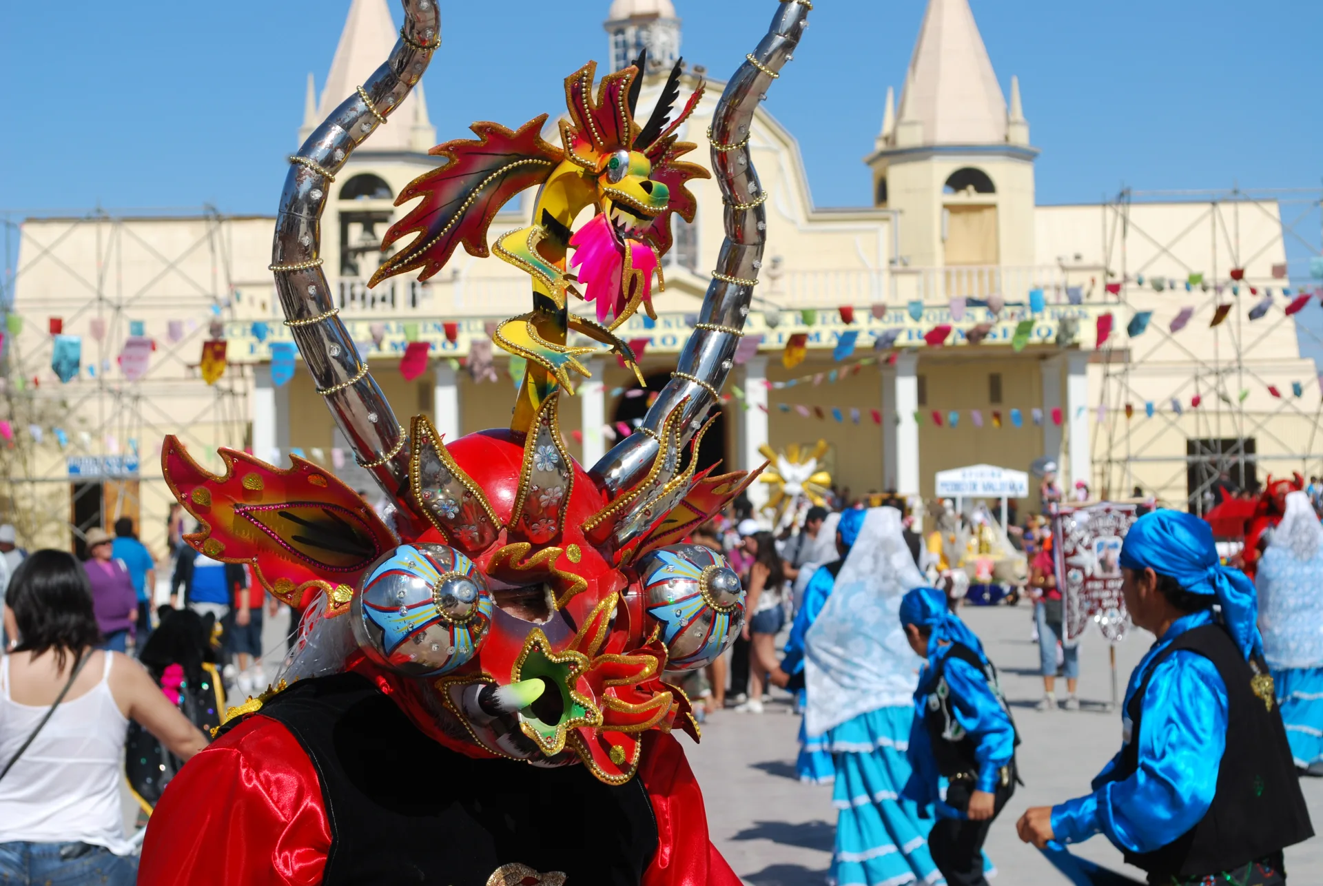 Máscara de diablada roja y dorada, con bailarines y el santuario de La Tirana al fondo