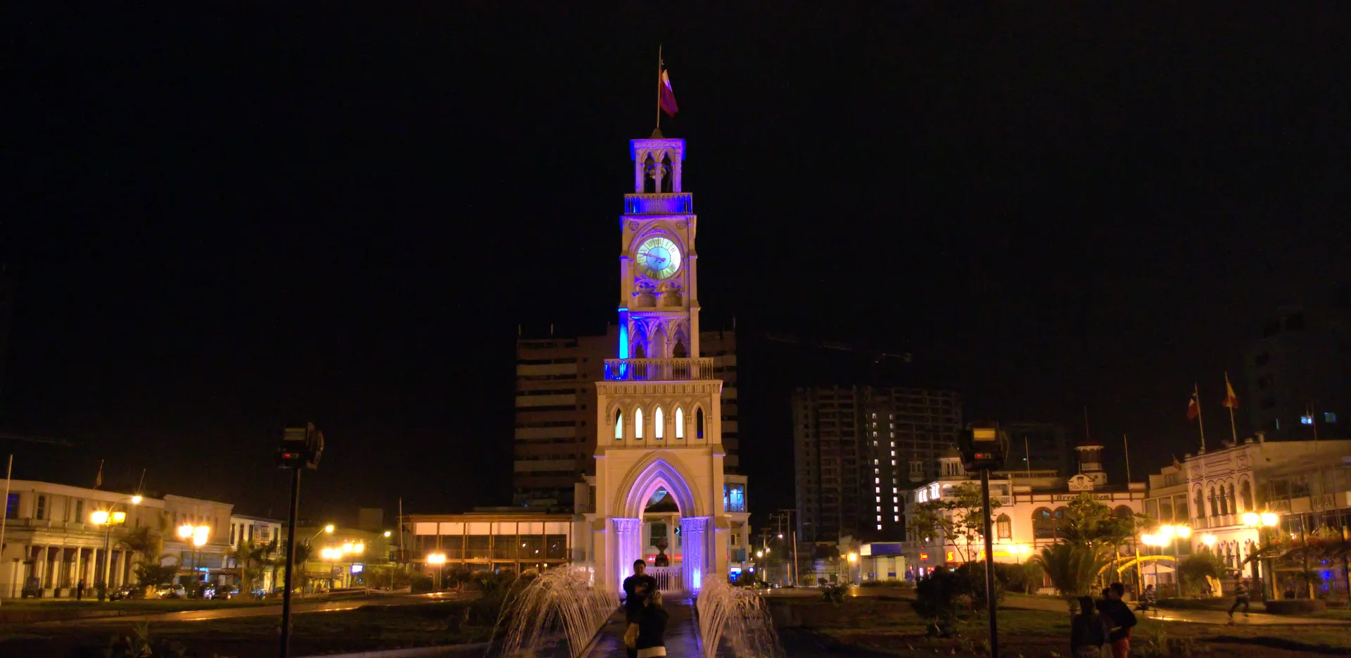 Torre del Reloj iluminada de noche, entre los edificios de la Plaza Arturo Prat de Iquique